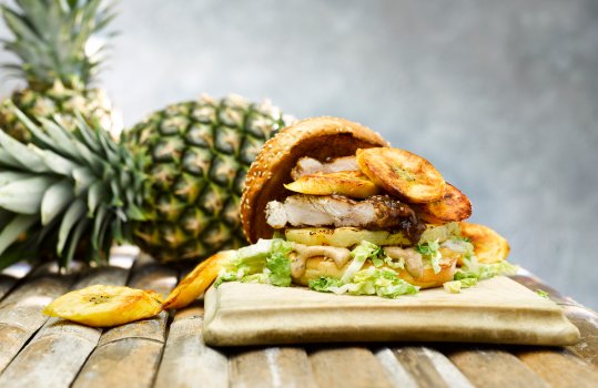 Food photograph of a Caribbean jerk spiced chicken thigh in a sandwich with grilled pineapple, roasted plantain, lettuce and jerk mayonnaise. Shot on a bamboo table with pineapples in the background