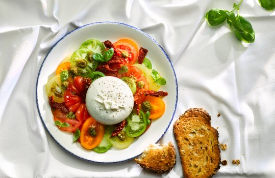 Aerial food photograph of a heritage tomato salad topped with a whole burrata, alongside slices of toasted sourdough bread