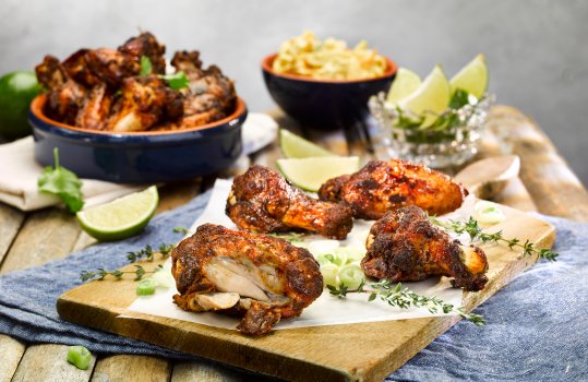 Food photograph close up of baked jerk chicken wings served on a wooden board alongside lime wedges, coleslaw and thyme sprigs