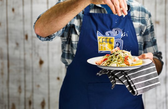 Food photograph of a plate of seafood linguine with lobster and crab, being topped with grated parmesan by a man wearing an apron