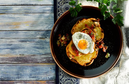 Food photograph of eggs Kejriwal, fried eggs with diced chilli and tomato, served on top of a crispy charred slice of cheese on sourdough toast, served with parsley sprigs on a painted blue tabletop with Indian patterned fabric