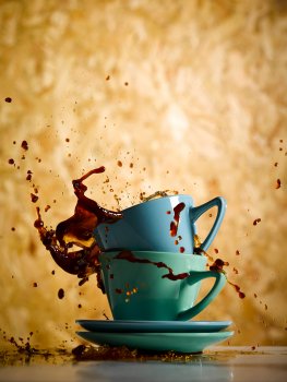 Drinks photograph of a stack of blue and green coffee cups, with coffee splashing out from the cups and onto the tabletop, with a wooden background