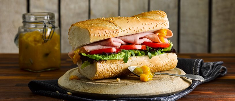 Food photograph of a ham salad baguette, a golden sesame seed baguette filled with fresh lettuce, sliced tomato and thick folded slices of ham, topped with picalilli, served on a wooden board with a blue tea towel on a dark wooden tabletop with a jar of homemade picalilli