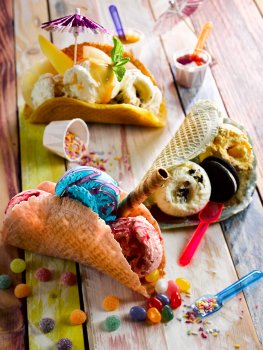 Food photograph of multi coloured ice cream tacos, scoops of bubblegum, strawberry, oreo, caramel and vanilla ice creams served in taco shells made from waffle batter, garnished with jelly tots, jelly beans, wafers, sprinkles, strawberry sauce, oreos and cocktail umbrellas; served on a multi coloured painted wooden table