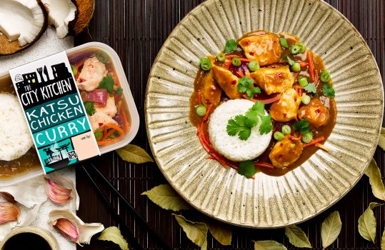 Aerial food photograph of a plated City Kitchen ready meal, chicken chunks in katsu sauce with steamed rice garnished with coriander and chopped spring onions, served on a bamboo mat alongside a packaged ready meal, a cracked coconut and scattered garlic cloves and dried bay leaves