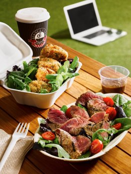 Food photograph of a bulgogi steak salad, seared rare steak with cherry tomatoes and leaves, along with a spiced chicken breast skewer served with salad, shot in an outdoor university setting on a wooden table with a laptop and a takeaway coffee in the background