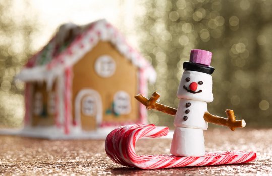 Food photography close up of a marshmallow snowman on a candy cane sleigh, marshmallows stacked and decorated to look like a snowman with pretzel arms and a liquorice hat, on a sled made from candy canes, shot in a festive setting on a glittering gold background with a gingerbread house