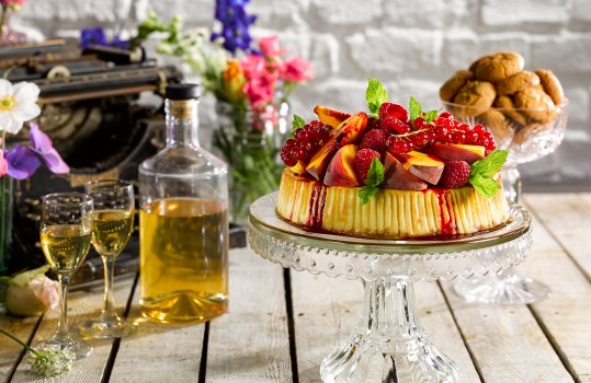 Food photograph of a summer peach melba cheesecake, a homemade peach cheesecake served on a vintage glass cake stand and topped with sliced peaches, raspberries and bunches of redcurrants, served in a vintage home style setting with an antique typewriter and flowers