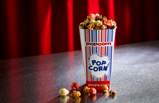 Food photograph of a cinema or comedy club portion of popcorn in a paper popcorn bag with kernels covered in various seasoning scattered around on a steel tabletop with a red curtain and spotlight in the background