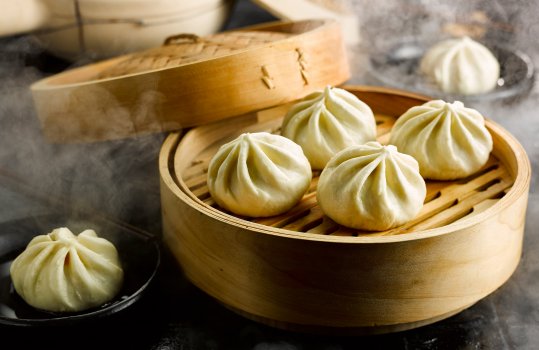 Food photograph of steamed char sui pork buns in a steam basket and being dipped into soy sauce in black ramekins, served on black shiny tiles with billowing clouds of steam