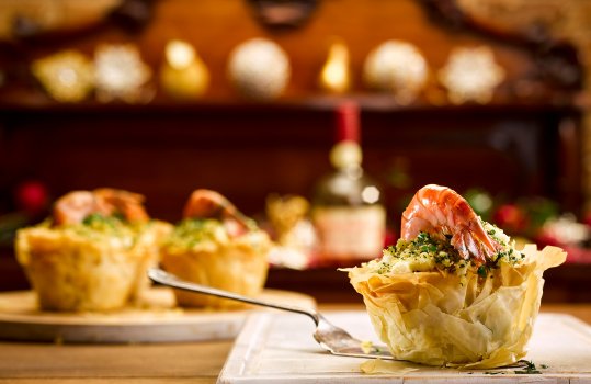 Food photograph close up of an individual fish pie, crisp filo pastry encasing fish pie mix and topped with herb breadcrumbs and a shiny king prawn in its shell. Shot in a festive home style setting on a white chopping board with an antique sideboard in the background with Christmas decorations and a vintage bottle of brandy