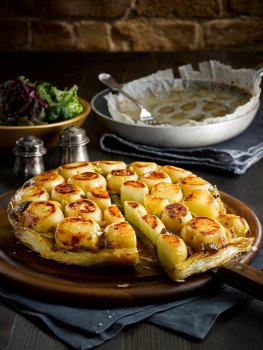 Food photograph of a baby potato tarte tatin, seared baby potatoes and caramelised onions topped with crisp puff pastry served on a wooden board with a bowl of salad and salt and pepper shakers, shot in a home style setting with the frying pan and parchment used to cook the tart in the background on a black wooden table and a brick wall