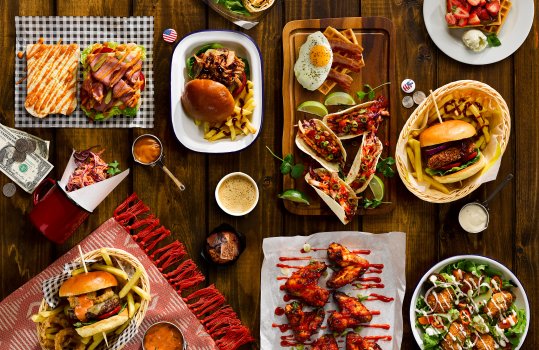 Aerial food photograph of an Americana sharing table, featuring a pastrami Rueben, cheeseburgers with onion rings and fries, waffles with bacon and eggs - and fresh strawberries and cream, pulled beef tacos with coriander and lime, grilled buffalo chicken wings and crispy battered popcorn chicken with salad, alongside a chocolate muffin with coffee, and various pots of slaw and dips, shot on a dark wooden setting