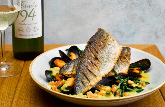 Food photograph of a restaurant main course, steamed mussels with samphire and butter sauce served underneath fillets of crispy pan fried sea bass, shot in a restaurant style setting on a wooden table with a bottle and glass of white wine, and a pale grey background