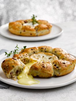 Food photograph of a baked camembert inside a cranberry tear and share bread, golden crusty bread studded with cranberries encasing a molten baked camembert which is spilling out where the bread has been torn, served on a white plate in a festive white and silver setting