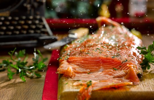 Food photograph of a whole side of homemade gin cured salmon, a whole side of cured glistening salmon topped with chopped dill and parsley and lemon juice being squeezed over the top, served sliced on a wooden board in a home style setting - on a wooden table with an antique typewriter in the background