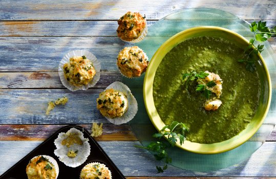 Aerial food photograph of homemade wild garlic soup served in a green bowl alongside homemade wild garlic and cheese mini muffins, shot in paper cupcake cases in a cupcake tray, shot on a painted blue and purple wooden background