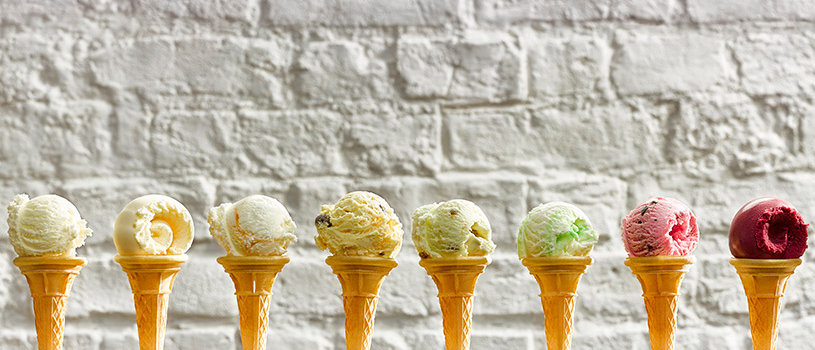 Food photograph of eight flavours of ice cream shown lined up in eight traditional style ice cream cones in front of a white painted brick background