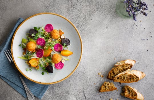 Aerial food photograph of a French restaurant starter, breadcrumbed and deep fried goats cheese bonbons, with roasted golden and regular beetroot, and slices of pickled beetroot garnished with micro herbs and toasted hazelnuts - shot on a grey stone background with a blue napkin, slices of toasted golden baguette and a small glass jar of lavender