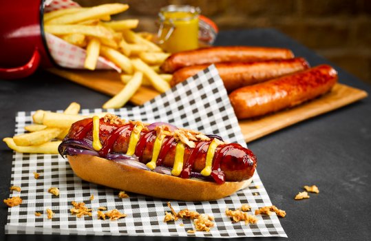 Food photograph of a homemade hot dog, a split top bun topped with a thick dark brown fried hot dog sausage with caramelised red onions, mustard, tomato ketchup and crispy fried onions, served on parchment paper with French fries and scattered crispy fried onions, shot on a dark home style setting on a slate tabletop with more hot dog sausages and a jar of mustard on a wooden bowl, and a red brick wall in the background