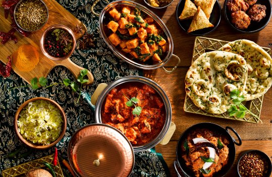 Aerial food photograph of an Indian spread, various curries including chicken tikka masala, butter paneer and lamb madras, served in a selection of metal balti dishes, alongside smaller dishes of pilau rice, chutneys, pakora and samosas - and a gold plate of garlic and coriander naan. Shot on a dark wooden table with Indian fabric and ramekins of spices, dried chillies and a coconut for decoration