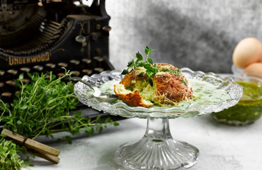 Food photograph of a twice baked pesto and cheese souffle with a golden brown exterior, served on a vintage glass cake stand with wild garlic veloute. Shot on a light grey setting with glass bowls of pesto and eggs, a bunch of thyme sprigs and an antique typewriter in the background