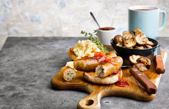 Food photograph of a homemade brunch, pan fried golden brown pork and leek sausages served with tomato chutney on a toasted English muffin alongside scrambled eggs and fried baby button mushrooms, shot on a wooden board in a light grey airy setting alongside a ramekin of tomato ketchup and a cup of coffee