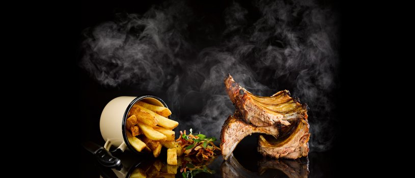 Food photograph of a fine dining grill restaurant main course, grilled and smoked pork ribs with a sticky glazed exterior served alongside a cup of skin on homemade chips and a pile of rainbow coleslaw, shot on a mirrored reflective tabletop on a black background, with wisps of smoke rising behind the stack of ribs