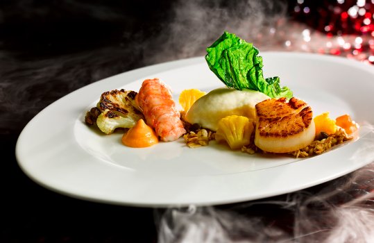 Food photograph of a fine dining fish course, a golden brown seared scallop and a steamed langoustine served with roasted, pickled and pureed cauliflower with red pepper sauce, crispy cabbage and toasted oats, shot on a white plate on an abstract red and black background with clouds of smoke