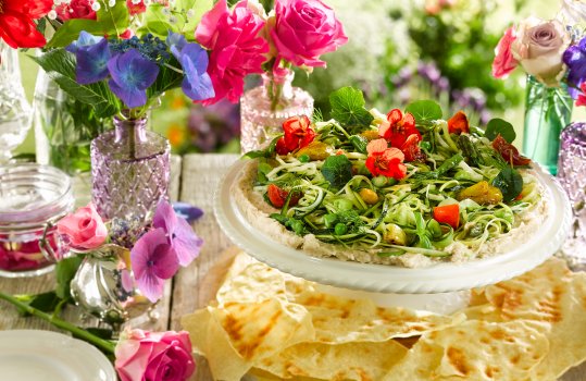 Food photograph of a homemade summer salad, thick homemade houmous topped with a salad of courgette, asparagus, garden peas, roasted peppers and roasted tomatoes, garnished with nasturtiums and served on a cake stand with crispy flatbread, shot in an outdoor setting on a picnic table with small glass vases of flowers