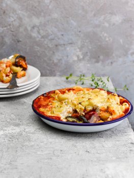 Food photograph of a vegan macaroni and Mediterranean roasted vegetable bake, an enamel dish filled with roasted aubergine, courgette, pepper and onion topped with tomato sauce and a layer of macaroni in cheese sauce. One spoonful has been removed from the dish and is served on a white plate in the background, shot on an abstract light grey setting