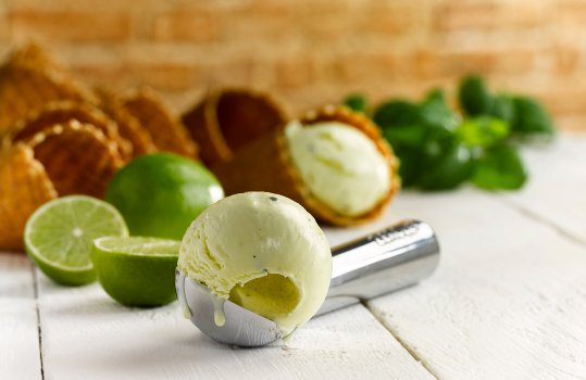 Close up food photograph of a scoop of pistachio flavoured ice cream shot inside an ice cream scoop on a painted white wooden table, shot alongside fresh limes, waffle cones and basil leaves