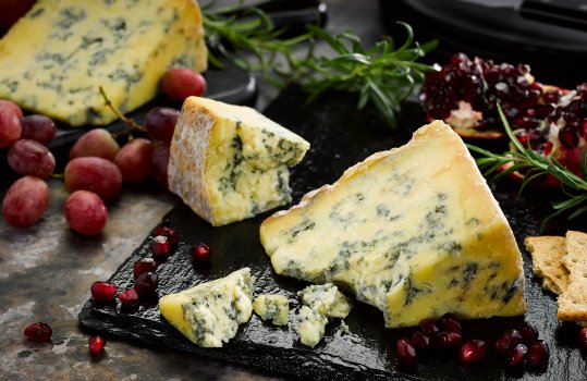 Food photograph of wedges of crumbly blue stilton cheese, served with grapes and pomegranate seeds on a slate slab on a dark setting