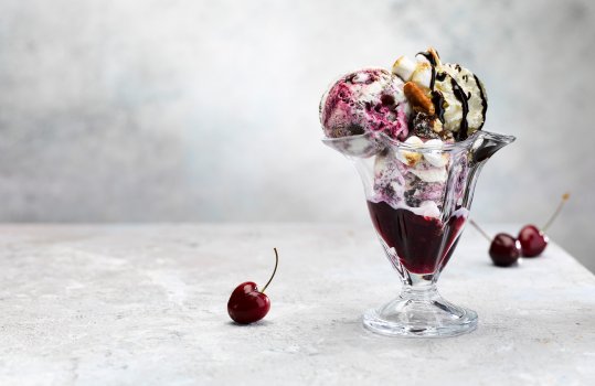 Food photograph of a black forest gateaux ice cream sundae, two scoops of black forest gateaux ice cream topped with toasted marshmallows, chocolate sauce and toasted pecans, served in a sundae glass with fresh black cherries on an abstract light grey set