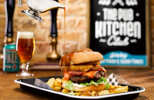 Food photograph of a cheeseburger in a sesame bun with onion rings, french fries, and onion gravy being poured over it from a metal jug.