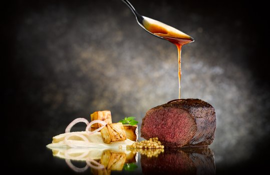 Food photograph of a fine dining main course, a juicy medium rare loin of venison with charred and pureed celeriac, and pickled onions and mustard seeds, shot on a reflective black abstract background with a spoon pouring jus over the venison