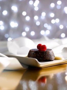 Food photograph of a miniature Christmas pudding, served on a white rectangular plate with custard and fresh cranberries. Shot on a reflective gold tabletop with white ribbon curled around the background, which also has sparkling Christmas lights