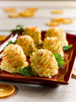 Food photograph of Duchess potatoes, a mix of mashed potato, butter, egg yolk, parsley and chilli which is poped using a star nozzle into mounds and then baked until golden and crisp, served on a dark red ceramic plate on a white painted wooden table with dried orange slices