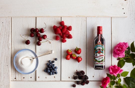 Aerial food photograph of ingredients to make a summer pudding, various fresh summer berries including blueberries, cherries, raspberries, strawberries and blackberries shot alongside a bottle of creme de cassis a bowl of white sugar, and pink roses on a white painted wooden background