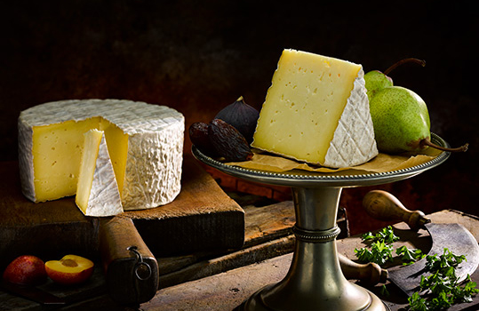 Food photograph of a whole wheel of Thelma’s traditional Caerffili cheese, the wheel is presented in the background on a chopping board with wedges having been cut from it and one wedge is served on an antique cake stand alongside fresh figs and pears, alongside a vintage herb cutter