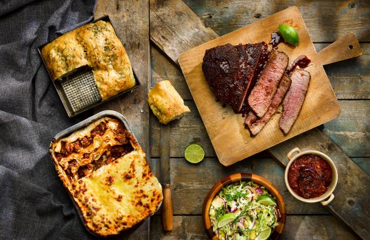 Aerial food photograph of a home made rich saucy lasagne al forno, a rustic home made focaccia topped with rosemary, and a sticky shiny glazed piece of barbecued beef, being sliced on a board alongside chilli sauce and a Brazilian pineapple coleslaw.