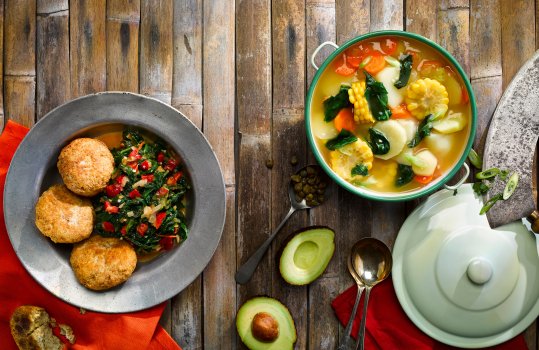 Aerial food photograph of spinach in the style of callaloo with fried dumplings, alongside a vegan ajiaco, a Colombian stew of corn and potatoes served with avocado, shot in vintage metal crockery, on a bamboo background