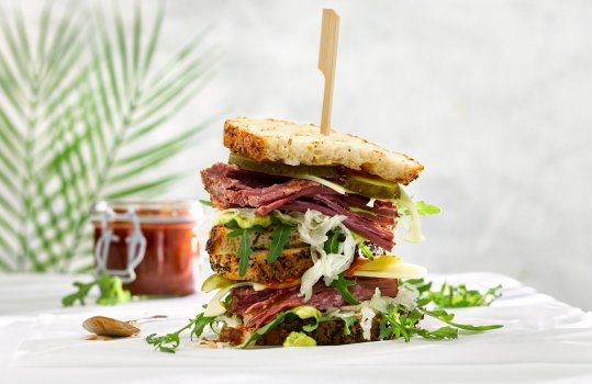 Food photograph close up of a stacked American salt beef sandwich, with sauerkraut, swiss cheese, rocket and pickles