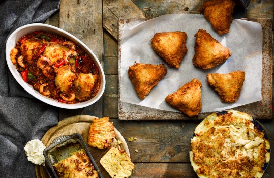 Aerial food photograph of a selection of Southern dishes, golden fried chicken quarters served on baking paper from a bucket, chicken and shrimp jambalaya served in a large ceramic dish, cheesy cornbread shot as a whole loaf with two slices cut out, and mac and cheese made with penne served in an enamel oven dish, all shot on a dark weathered wooden background