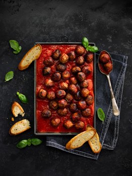 Aerial food photograph of a tray of meat free meatballs in a tray of rich tomato sauce, alongside toasted slices of ciabatta and sprigs of basil, shot on a black slate background