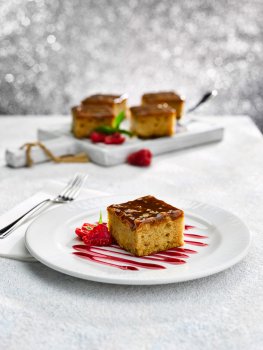 Food photograph close up of a Christmas dessert, a spiced apple cake topped with caramel sauce, served on a white plate with the rest of the tray cake on a board in the background, shot against a glittering background