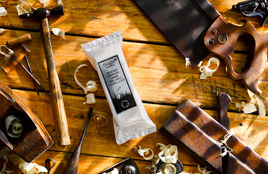 Aerial food photograph of four packs of Dragon Handcrafted cheddar cheese, surrounded on a wooden tabletop by vintage woodworking tools and wood shavings