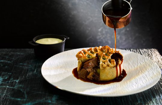 Food photograph close up of a fine dining main course, an individual beef pie with golden flaky pastry and large pieces of braised beef within, studded with thyme leaves. Served on a white textured plate with a small ramekin of pomme puree, and gravy being poured over the pie