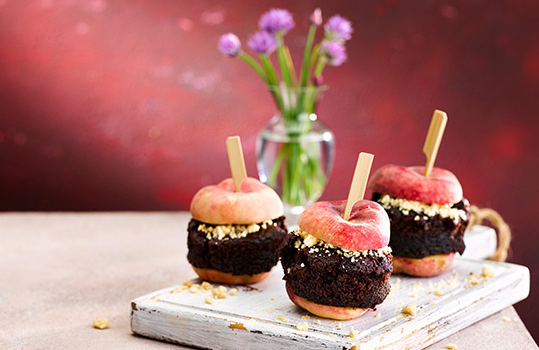 Food photograph of a vegan recipe for ‘doughnut peach burgers’, a brownie ‘patty’ in between two halves of a flat peach which are serving as ‘buns’. Shot on a dark red setting with a white serving board