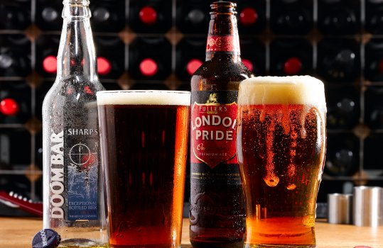 Drinks photograph of two pints of dark foamy British beers, shot having been poured into pint glasses with the bottles shown behind, shot on a wooden table with a bottle rack in the background